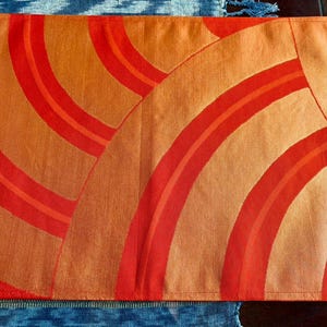 May include: Orange and red rectangular placemat with a repeating curved stripe design. The placemat has a textured appearance and is bordered by a stitched edge. The design features varying shades of orange and red.