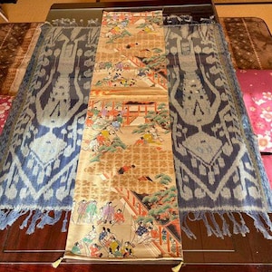 以下が含まれることがあります： 伝統的な衣装を着た人物、建物、木々を描いた、歴史的なシーンの詳細なイラストが施された装飾用テキスタイル。 テキスタイルは、濃い木製の表面に配置され、青と白の模様の生地で挟まれています。