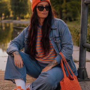 Könnte beinhalten: Eine orangefarbene Strickmütze mit einem schwarzen Etikett mit der Aufschrift Rock aus Liebe, eine blaue Jeansjacke, ein orange-blau gestreiftes Langarmshirt, eine blaue Jeans, weiße Socken, orangefarbene Sneaker und eine orangefarbene Häkeltasche.