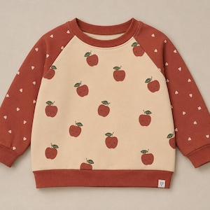 Könnte beinhalten: Ein langärmeliges Sweatshirt mit einem cremefarbenen Mittelteil, das mit einem sich wiederholenden Muster aus kleinen roten Äpfeln bedruckt ist. Die Ärmel und der Saum sind rostrot, wobei die Ärmel mit winzigen weißen Herzen verziert sind.