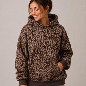 Peut inclure: Un sweat à capuche à manches longues avec un motif léopard marron et noir sur l'ensemble. Le vêtement comprend une grande poche kangourou à l'avant, une capuche et des bords-côtes marron foncé aux manches et à la base pour la structure.