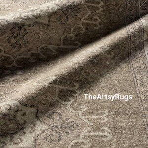 May include: Close-up of a beige and gray area rug with an intricate, traditional pattern. The rug has a distressed look with a border design and the text "TheArtsyRugs" in the bottom right corner.