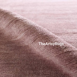Puede incluir: Primer plano de una alfombra de color rosa polvoriento con una superficie texturizada. La alfombra est&aacute; enrollada, revelando el tejido y el pelo. El texto "TheArtsyRugs" es visible en la esquina inferior derecha.