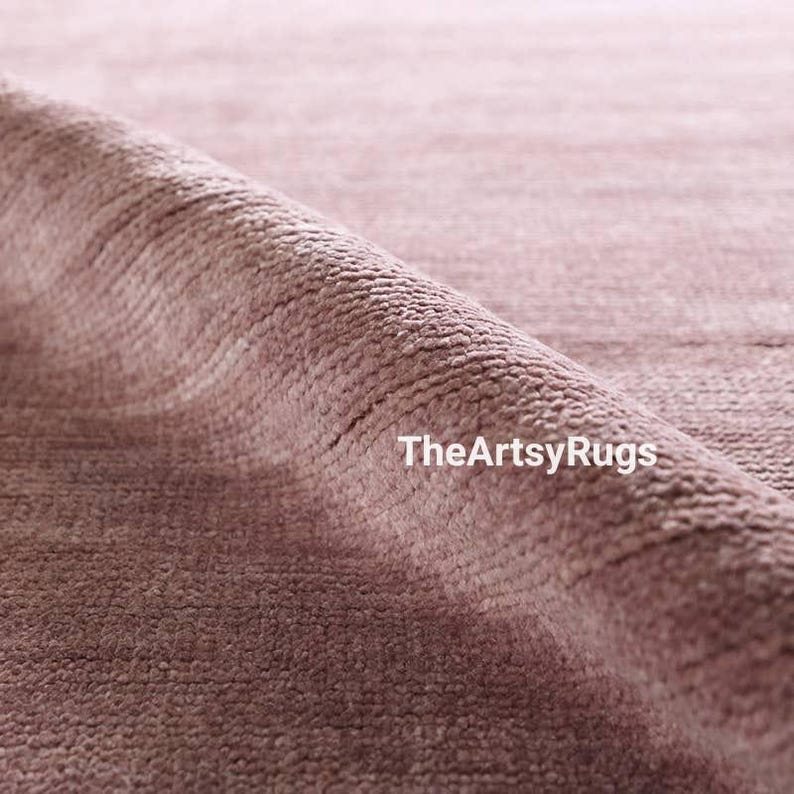 Puede incluir: Primer plano de una alfombra de color rosa polvoriento con una superficie texturizada. La alfombra est&aacute; enrollada, revelando el tejido y el pelo. El texto "TheArtsyRugs" es visible en la esquina inferior derecha.