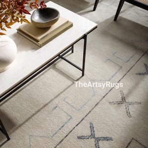 May include: A neutral-toned rug with a geometric design featuring light blue and gray accents. The rug is paired with a white marble coffee table with black metal legs. A decorative bowl and books are on the table.