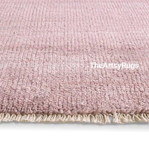 Puede incluir: Primer plano de una alfombra de color rosa empolvado con un tejido texturizado. La alfombra tiene una franja a lo largo del borde inferior. El texto "TheArtsyRugs" es visible en la superficie de la alfombra.