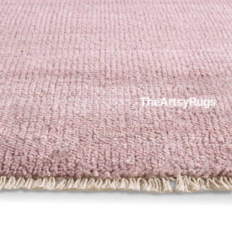 Puede incluir: Primer plano de una alfombra de color rosa empolvado con un tejido texturizado. La alfombra tiene una franja a lo largo del borde inferior. El texto "TheArtsyRugs" es visible en la superficie de la alfombra.