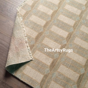 May include: A rectangular rug with a geometric pattern in beige, light blue, and tan. The rug's design features rectangular and circular shapes. The rug is partially rolled up, revealing its underside and fringe. The text "TheArtsyRugs" is visible.