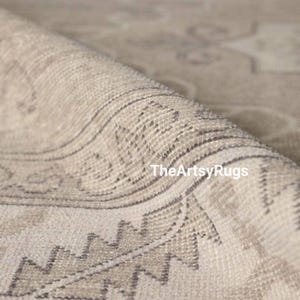 May include: Close-up of a rolled-up beige rug with a detailed geometric pattern. The rug features a combination of light beige and gray tones, with a visible texture. The text "TheArtsyRugs" is visible on the rug.