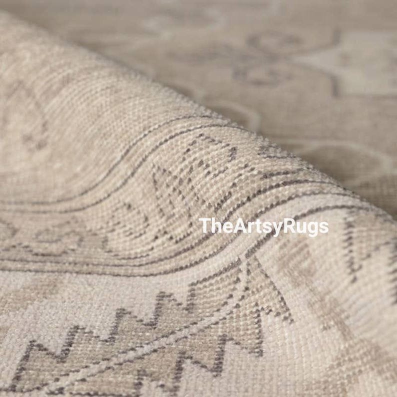 May include: Close-up of a rolled-up beige rug with a detailed geometric pattern. The rug features a combination of light beige and gray tones, with a visible texture. The text "TheArtsyRugs" is visible on the rug.