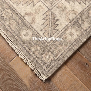 May include: Close-up of a corner of a beige and gray patterned rug with a geometric design. The rug has a fringe edge and the text "TheArtsyRugs" is visible. The rug is on a wooden floor.