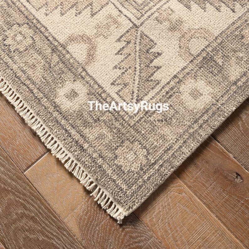 May include: Close-up of a corner of a beige and gray patterned rug with a geometric design. The rug has a fringe edge and the text "TheArtsyRugs" is visible. The rug is on a wooden floor.