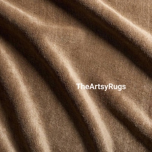 May include: Close-up of a textured, brown fabric with diagonal folds. The fabric has a soft, velvety appearance. The text "TheArtsyRugs" is printed in white across the bottom of the image. This fabric is suitable for rugs.