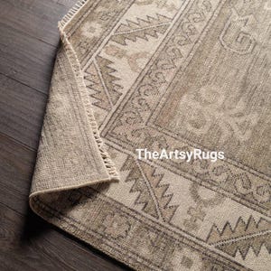 May include: A rolled-up, rectangular area rug with a geometric pattern in shades of beige and brown. The rug has a fringe on one edge and the text "TheArtsyRugs" is visible.