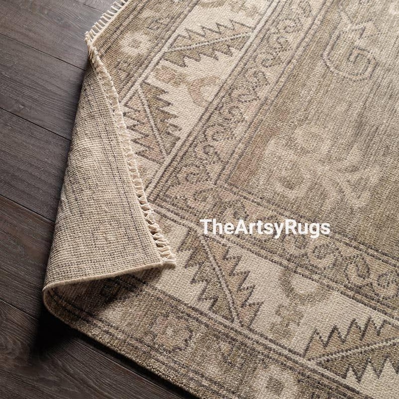 May include: A rolled-up, rectangular area rug with a geometric pattern in shades of beige and brown. The rug has a fringe on one edge and the text "TheArtsyRugs" is visible.