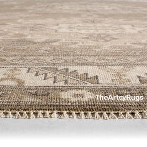 May include: Close-up of a beige and brown patterned rug with a fringe edge. The rug features a detailed geometric design and the text "TheArtsyRugs" is visible in the bottom right corner. The rug is made of a woven material.