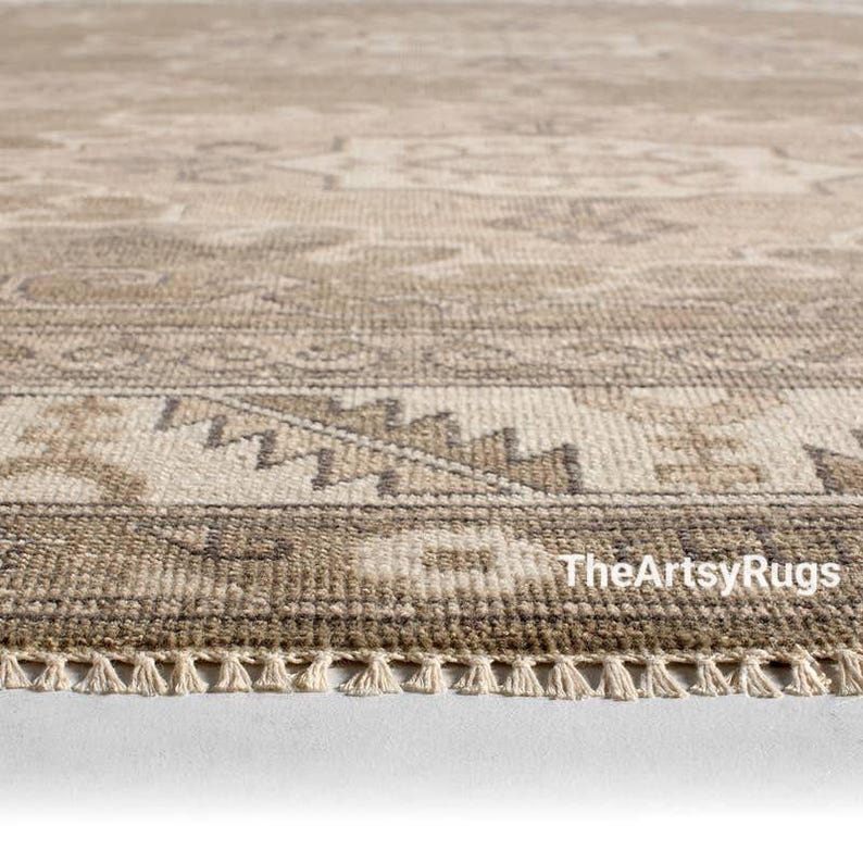 May include: Close-up of a beige and brown patterned rug with a fringe edge. The rug features a detailed geometric design and the text "TheArtsyRugs" is visible in the bottom right corner. The rug is made of a woven material.