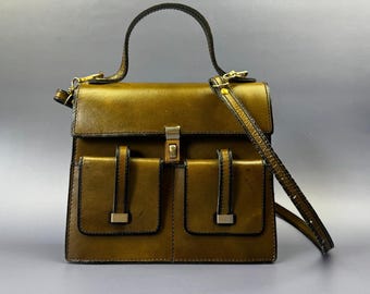 Puede incluir: Un bolso de mano estructurado de cuero verde oliva con asa superior y correa de hombro desmontable. El bolso presenta un cierre de solapa rectangular con un broche metálico central y dos bolsillos exteriores frontales, cada uno asegurado con una tira vertical.