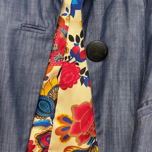 May include: A blue blazer with a single black button featuring an embossed logo. The interior lining is visible, showcasing a vibrant, multi-colored floral and paisley pattern in shades of red, yellow, blue, and orange on a cream background.