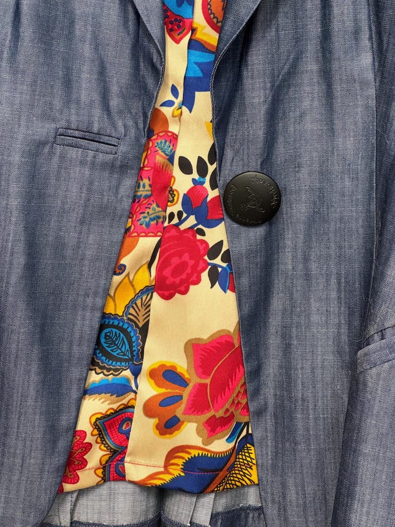 May include: A blue blazer with a single black button featuring an embossed logo. The interior lining is visible, showcasing a vibrant, multi-colored floral and paisley pattern in shades of red, yellow, blue, and orange on a cream background.