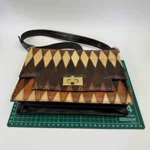 May include: A vintage handbag with a geometric pattern in shades of brown, tan, and black. The bag features a gold-colored clasp and a dark brown shoulder strap. The bag is sitting on a green cutting mat.