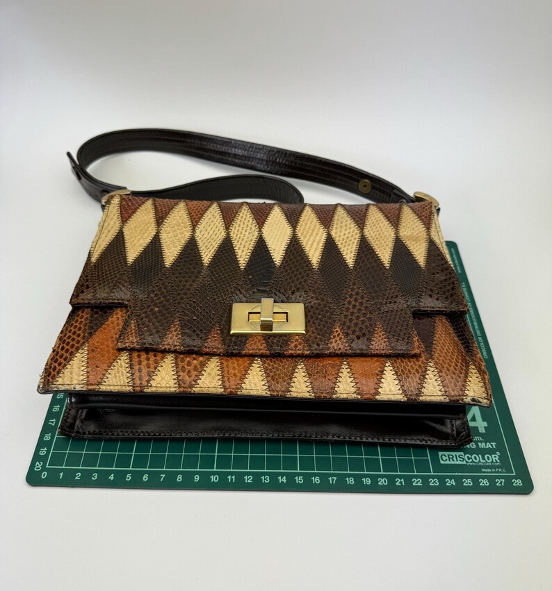 May include: A vintage handbag with a geometric pattern in shades of brown, tan, and black. The bag features a gold-colored clasp and a dark brown shoulder strap. The bag is sitting on a green cutting mat.