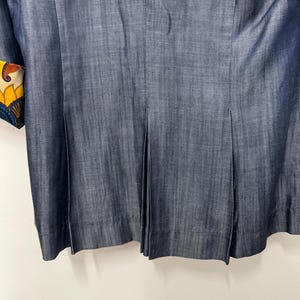 May include: A dark blue denim blazer featuring vertical pleats along the lower back section. The sleeve cuff displays a vibrant, multi-colored floral and paisley pattern in shades of yellow, orange, blue, and white, contrasting with the solid fabric.
