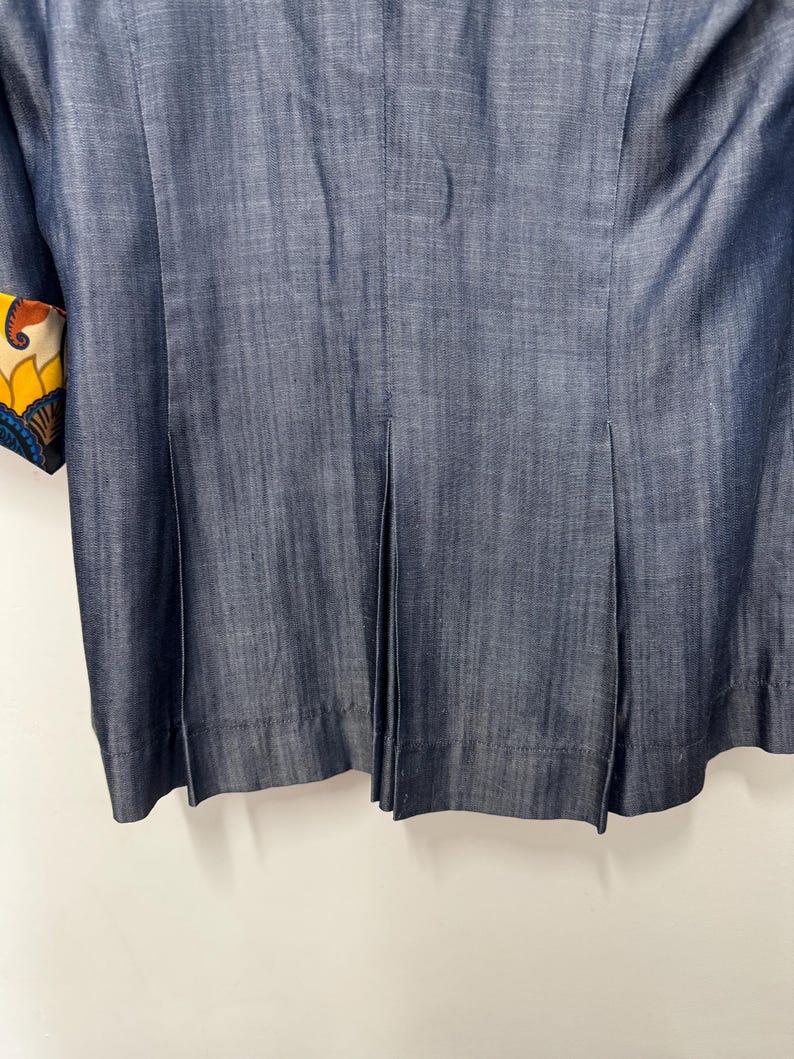 May include: A dark blue denim blazer featuring vertical pleats along the lower back section. The sleeve cuff displays a vibrant, multi-colored floral and paisley pattern in shades of yellow, orange, blue, and white, contrasting with the solid fabric.