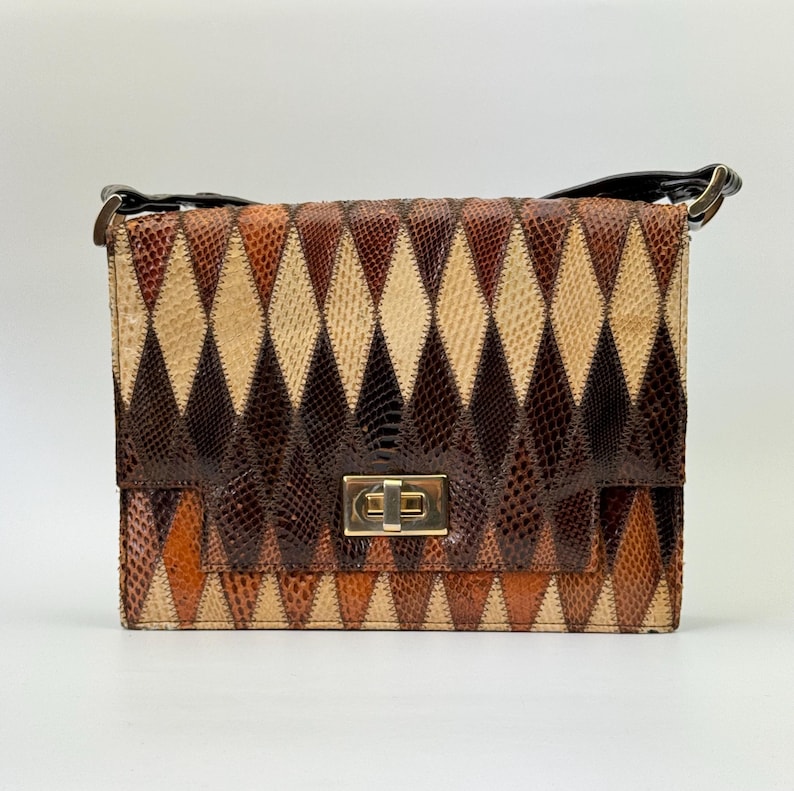 May include: A rectangular handbag with a geometric diamond pattern in shades of brown, tan, and orange. The bag features a gold-colored clasp and a black handle. The design resembles snakeskin.