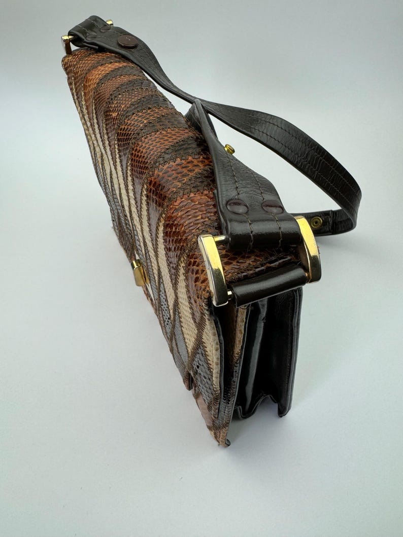 May include: A vintage handbag with a snakeskin pattern in shades of brown, tan, and black. The bag features a black leather strap and gold-colored hardware. The bag is rectangular and has a clasp closure.