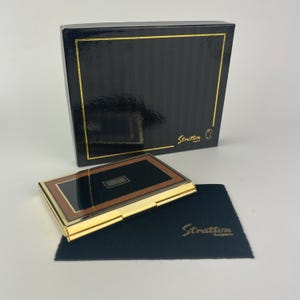 May include: A rectangular Stratton England compact case featuring a black and brown border with a gold-tone metal frame. The set includes a black presentation box with gold trim and a black fabric pouch, both displaying the Stratton England logo.