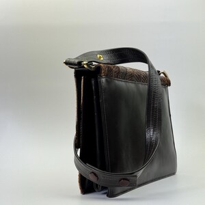 May include: A vintage black leather handbag with a brown patterned top flap and handle. The bag features a long, adjustable strap and a gold-toned clasp. The bag has a structured design and a classic, timeless aesthetic.