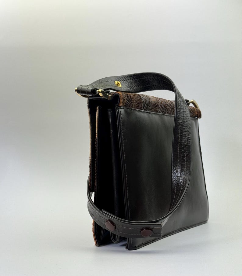 May include: A vintage black leather handbag with a brown patterned top flap and handle. The bag features a long, adjustable strap and a gold-toned clasp. The bag has a structured design and a classic, timeless aesthetic.