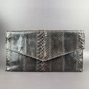 Könnte beinhalten: Eine rechteckige Clutch im Umschlag-Stil mit einem dunkelbraunen und schwarzen Schlangenledermuster und einer ausgeprägten Schuppenstruktur. Die Tasche hat einen spitzen Klappenverschluss, der sich über die Vorderseite erstreckt.
