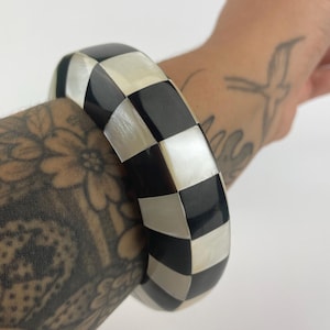 Può includere: Un bracciale rigido largo caratterizzato da un motivo a scacchiera ripetuto. Il design è composto da tessere quadrate alternate in nero solido e madreperla bianca iridescente, creando un look geometrico ad alto contrasto lungo tutto il cerchio.