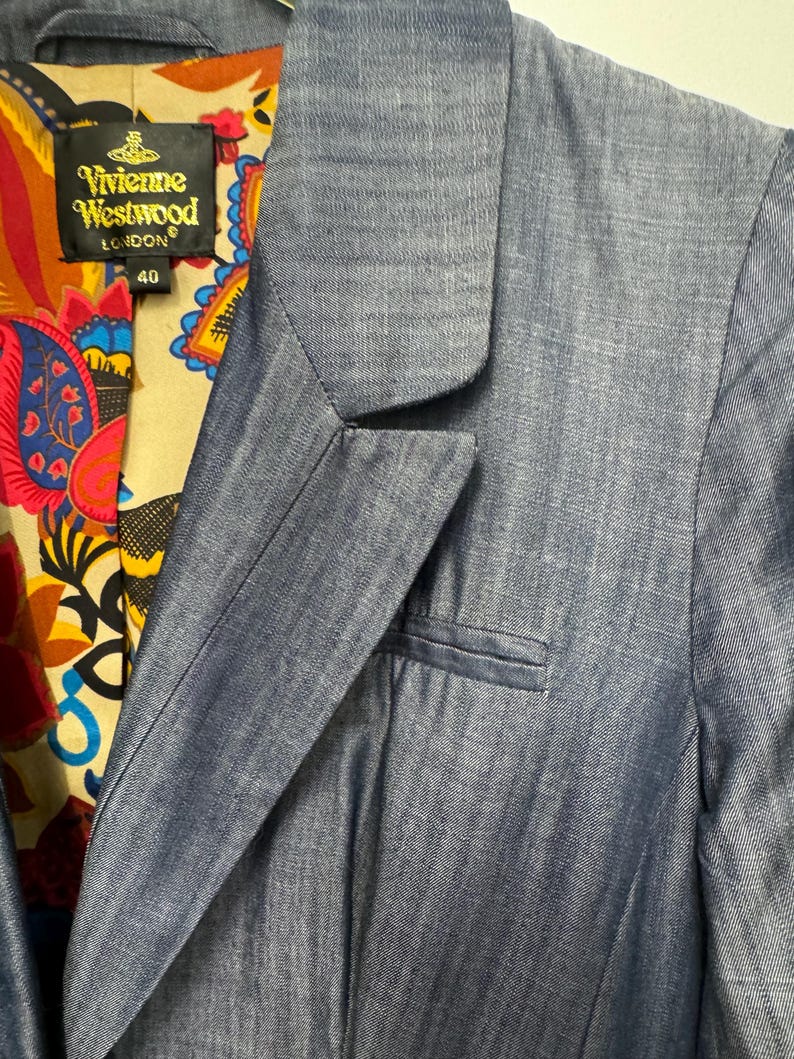 May include: A blue blazer with a notched lapel and a welt pocket on the chest. The interior lining features a colorful paisley pattern in red, yellow, and blue. A black label with gold text reads Vivienne Westwood London and the number 40.