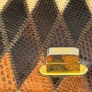 May include: Close-up of a handbag featuring a geometric pattern in shades of brown, tan, and cream. The bag's surface has a textured, reptile-like appearance, with a gold-colored clasp. The stitching is visible, adding to the bag's design.