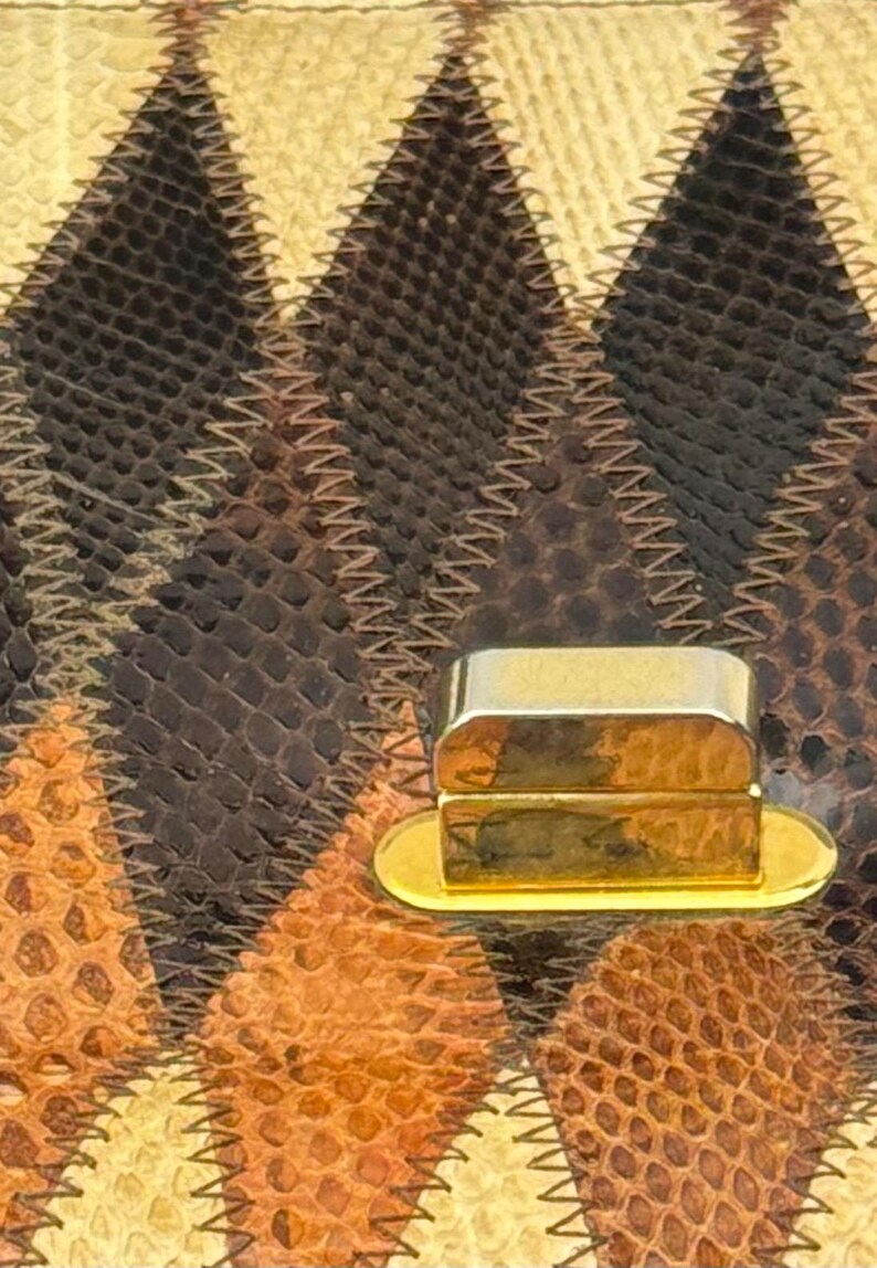 May include: Close-up of a handbag featuring a geometric pattern in shades of brown, tan, and cream. The bag's surface has a textured, reptile-like appearance, with a gold-colored clasp. The stitching is visible, adding to the bag's design.