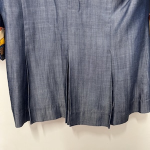 May include: A dark blue denim-textured jacket featuring a vertical seam down the center back and two symmetrical pleats extending from the waist to the hem. The fabric displays a subtle cross-hatch weave pattern with a visible hemline at the bottom.