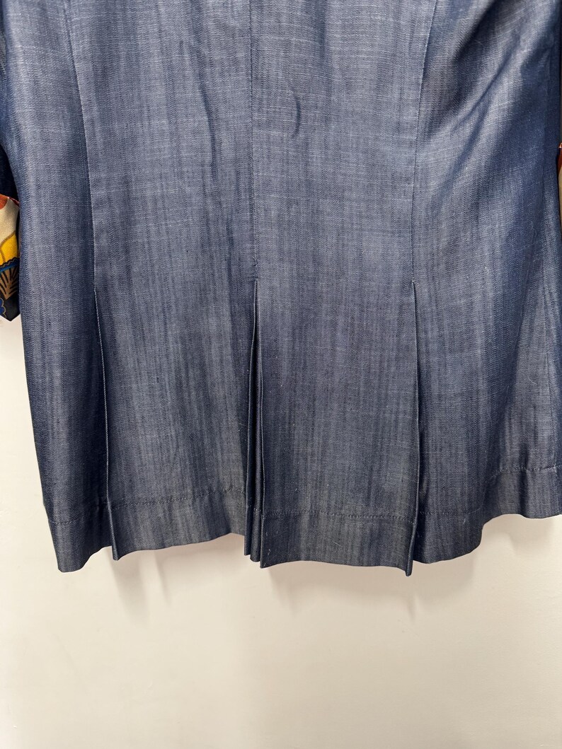 May include: A dark blue denim-textured jacket featuring a vertical seam down the center back and two symmetrical pleats extending from the waist to the hem. The fabric displays a subtle cross-hatch weave pattern with a visible hemline at the bottom.