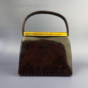 Peut inclure: Un sac à main structuré avec un extérieur texturé en peau de serpent brun foncé brillant. Le sac possède une anse arquée unique et une fermeture à cadre métallique rectangulaire dorée qui s'étend sur toute la largeur de l'ouverture.