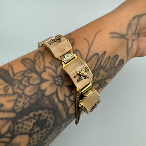 Könnte beinhalten: Ein goldfarbenes Gliederarmband mit rechteckigen, durchscheinenden, cremeweißen Platten. Jede Platte ist mit einer kleinen, metallischen Figur einer Person verziert, die Gitarre spielt. Die Glieder sind durch verzierte, filigrane Goldverbindungen verbunden.