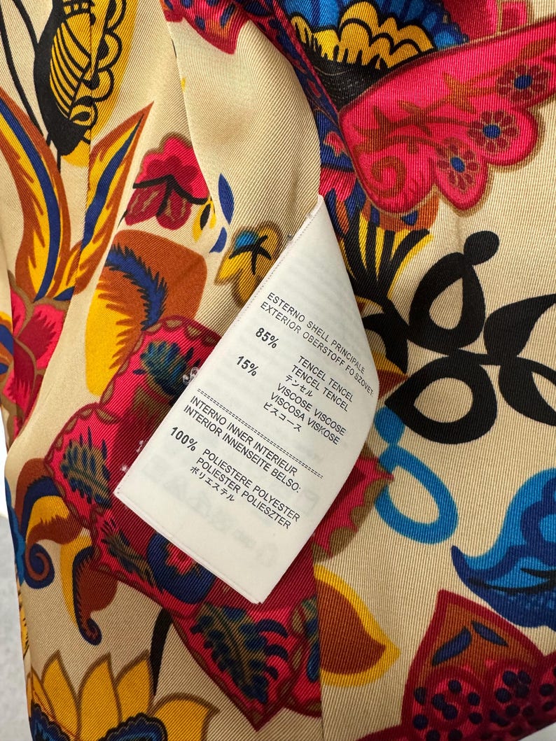 May include: A garment interior featuring a vibrant floral pattern in shades of red, yellow, blue, and black on a beige background. A white care label is attached, listing 85% Tencel, 15% Viscose for the shell, and 100% Polyester for the inner lining.