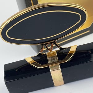 Può includere: Un pezzo decorativo nero di forma ovale con un bordo dorato, fissato a una clip in metallo dorato che si aggancia a un oggetto nero rettangolare. Il design presenta una finitura liscia e lucida con linee pulite e un accento dorato.