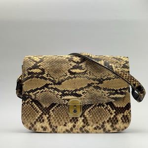 May include: A rectangular handbag with a snakeskin pattern in shades of brown and tan. The bag has a flap closure with a gold-colored clasp and a black shoulder strap. The bag is made of a textured material.