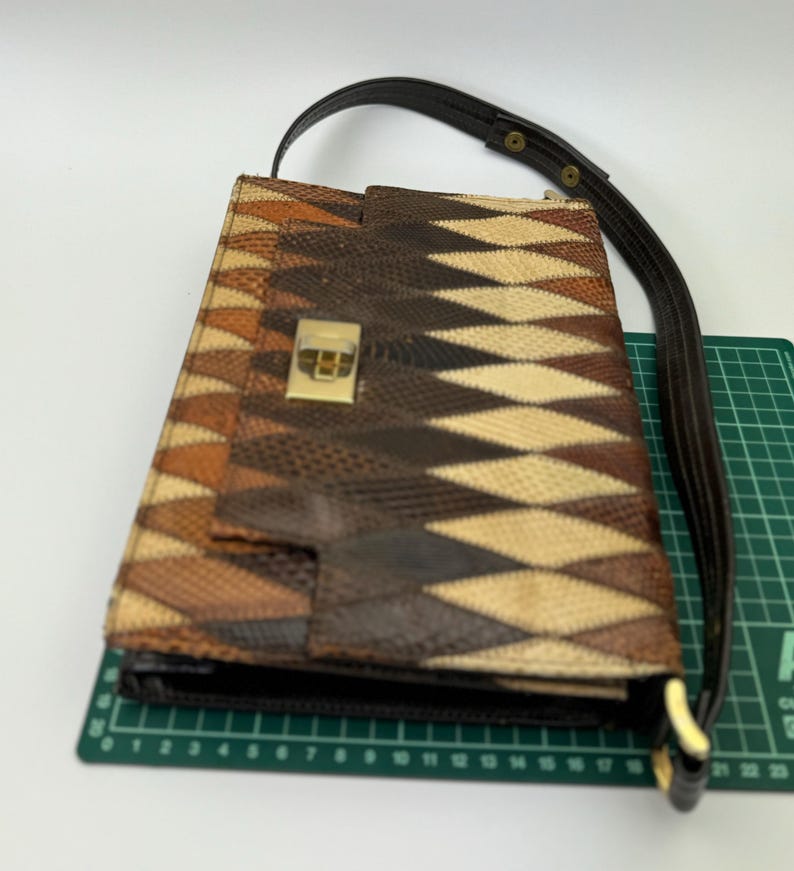 May include: A brown and tan patterned handbag with a dark brown strap. The bag features a geometric design and a gold-colored clasp. The strap is attached with gold-colored hardware.