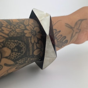 Peut inclure: Un bracelet jonc géométrique composé de panneaux alternés en matériau noir poli et en nacre blanche irisée. Le bracelet présente un design facetté et angulaire avec des bords nets et une large bande structurée qui entoure le poignet.