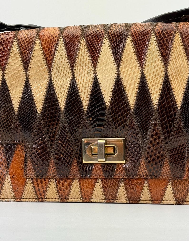 May include: A rectangular handbag with a geometric diamond pattern in shades of brown, tan, and black. The bag features a gold-colored clasp and a black strap. The design resembles a patchwork or quilt, with each diamond segment stitched together.