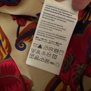 May include: A white rectangular garment care label with multi-language text and laundry symbols, including instructions for cold machine washing, no bleaching, no tumble drying, and low heat ironing, attached to a fabric with a colorful floral pattern.