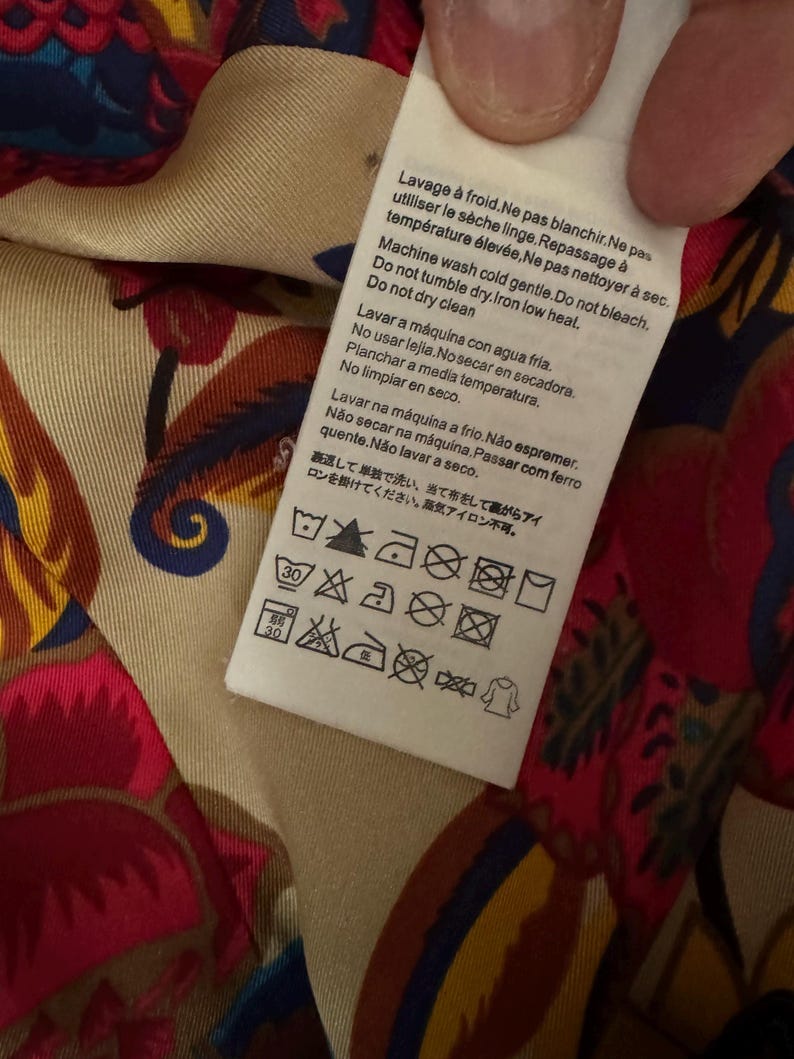 May include: A white rectangular garment care label with multi-language text and laundry symbols, including instructions for cold machine washing, no bleaching, no tumble drying, and low heat ironing, attached to a fabric with a colorful floral pattern.